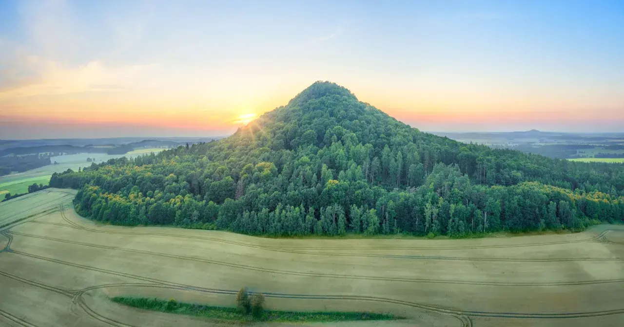 Wschód słońca nad krainą wygasłych wulkanów. Zielone wzgórze porośnięte lasem, pola uprawne i łagodnie pofałdowany krajobraz w oddali.