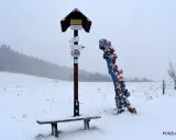 Zimowy krajobraz z ławką i drogowskazem turystycznym. Obok kolorowa rzeźba, być może symbolizująca skopiec szczyt.