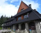 Kamienny schron turystyczny pod gorącymi szczytami, z drewnianą przybudówką i czarnym dachem, otoczony lasem świerkowym.