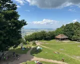 Ludzie odpoczywają na trawie i spacerują po szlakach na górze Ślęża. W oddali widać rozległą panoramę.