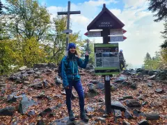 Kobieta na szczycie Łysicy, 614 m n.p.m., z drewnianym krzyżem i drogowskazem szlaku. Jesienne liście i kamienie wokół.