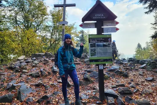 Kobieta na szczycie Łysicy, 614 m n.p.m., z drewnianym krzyżem i drogowskazem szlaku. Jesienne liście i kamienie wokół.