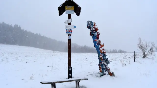 Zimowy krajobraz z ławką i drogowskazem turystycznym. Obok kolorowa rzeźba, być może symbolizująca skopiec szczyt.