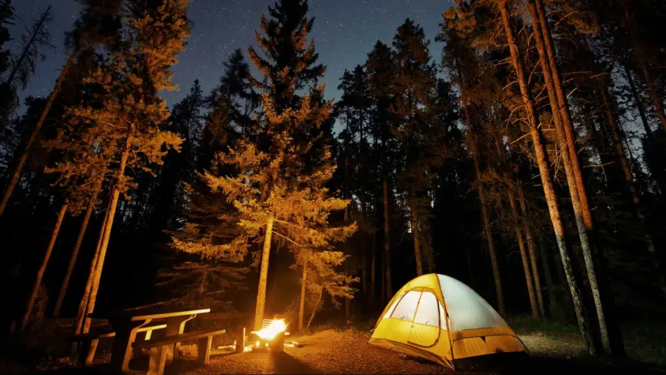 Nocny **pole namiotowe z widokiem na góry**. Rozświetlony namiot, ognisko i ławka wśród drzew pod rozgwieżdżonym niebem.