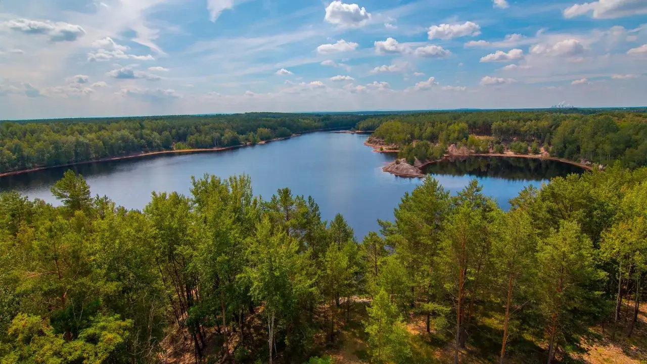 Malownicze jezioro w otoczeniu lasów, część geoparku Łuk Mużakowa. Błękitne niebo z chmurami odbija się w spokojnej tafli wody.