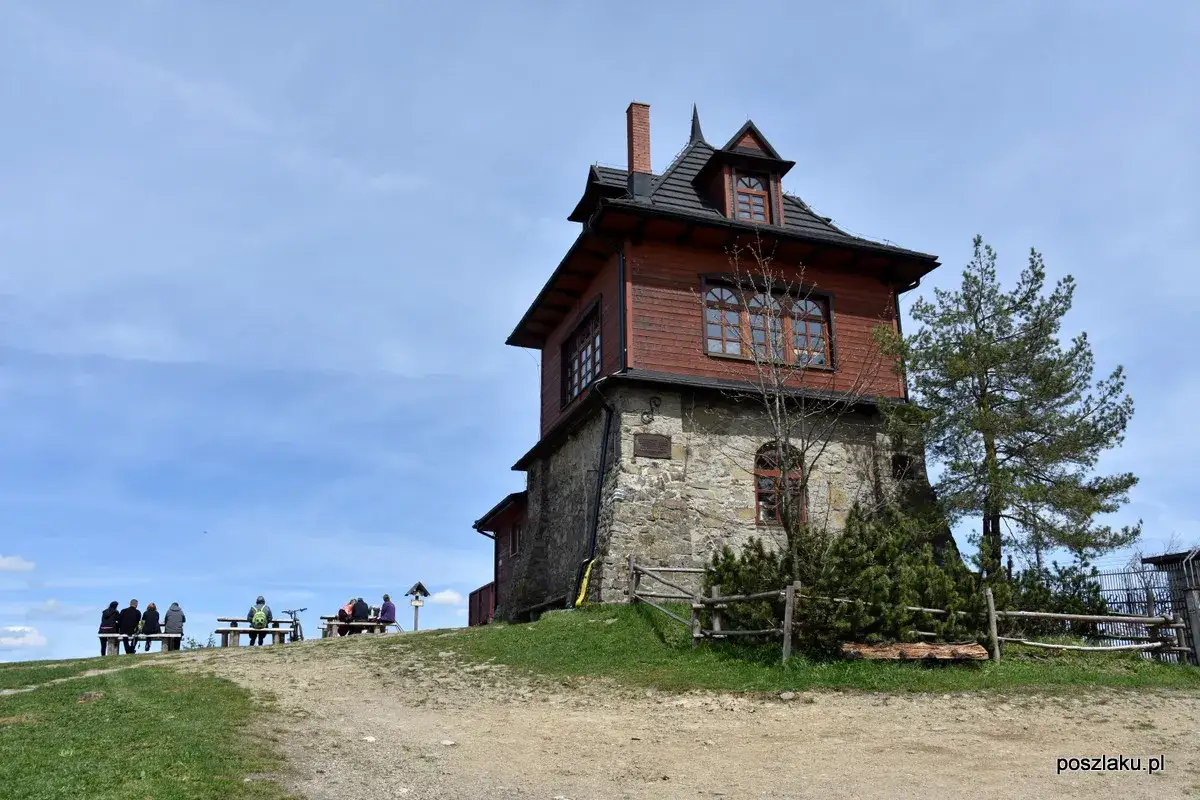 Drewniane schronisko na Luboniu Wielkim, z kamiennym fundamentem. Ludzie odpoczywają na ławkach na tle nieba.